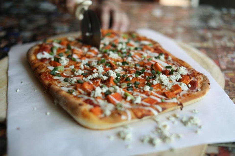 sicilian crust pizza with red sauce, mozzarella, chicken, buffalo sauce, green onions, drizzled with ranch. Pizza is being cut iwht a pizza cutter.